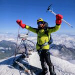 A triumphant mountaineer wearing a bright yellow-green hooded winter jacket, black snow pants, blue goggles, yellow helmet, and oversized red insulated mittens stands atop a snowy mountain summit