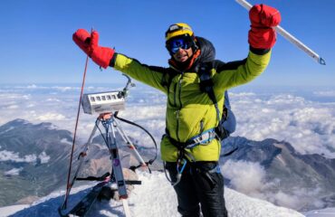 A triumphant mountaineer wearing a bright yellow-green hooded winter jacket, black snow pants, blue goggles, yellow helmet, and oversized red insulated mittens stands atop a snowy mountain summit