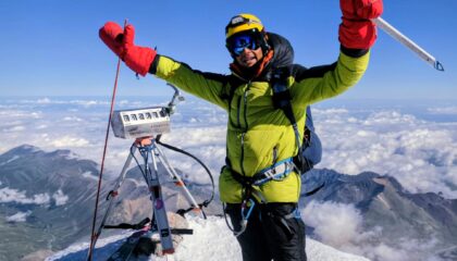 A triumphant mountaineer wearing a bright yellow-green hooded winter jacket, black snow pants, blue goggles, yellow helmet, and oversized red insulated mittens stands atop a snowy mountain summit