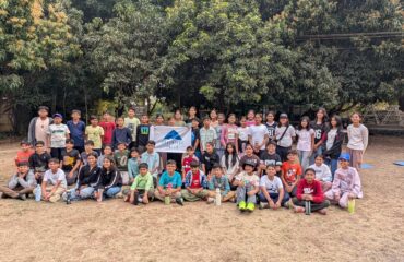 A large group of diverse children and teenagers posing together for a commemorative outdoor group photo on a sandy ground