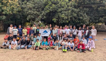 A large group of diverse children and teenagers posing together for a commemorative outdoor group photo on a sandy ground
